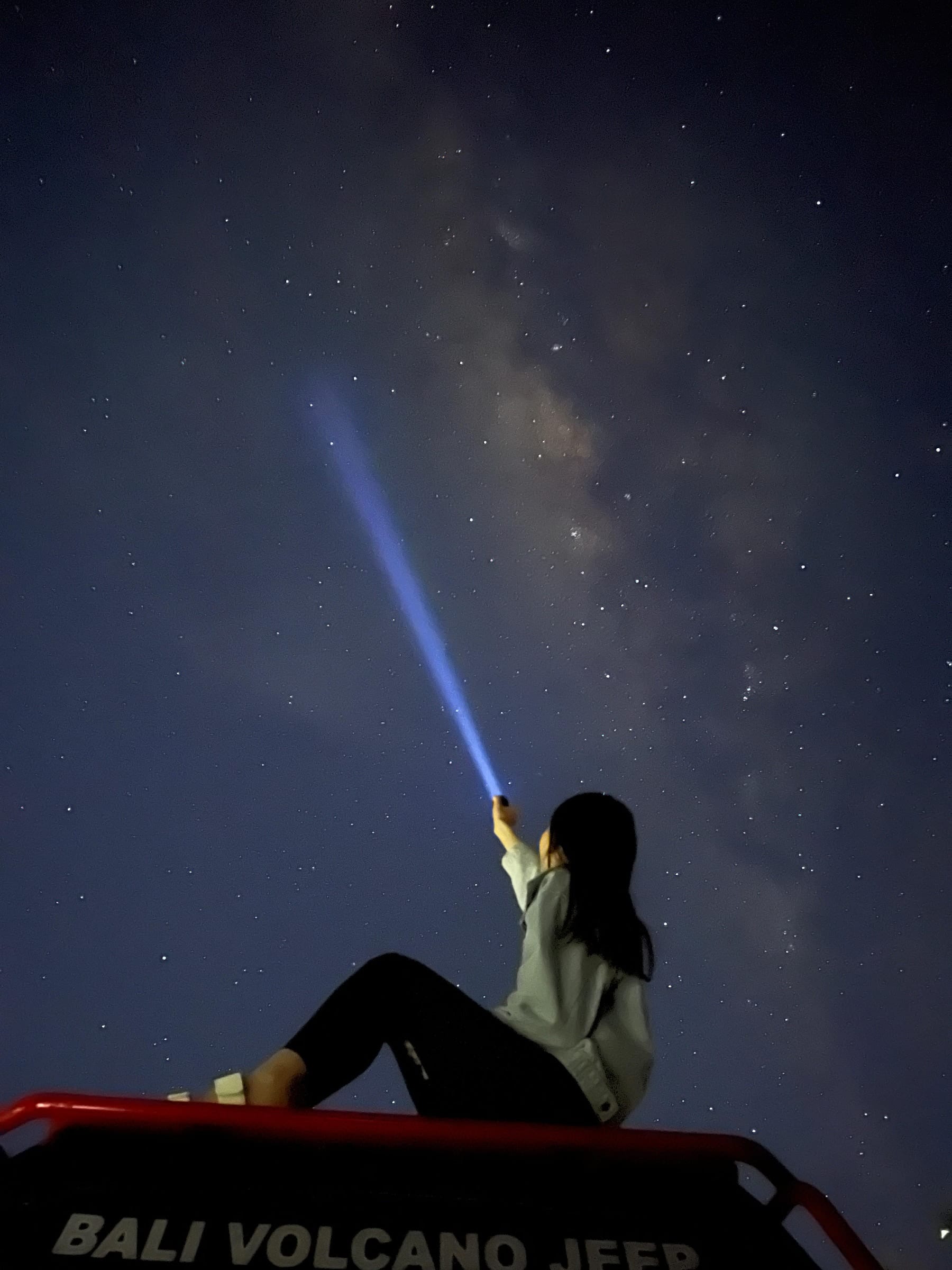 A guest sitting on a jeep roof before dawn, pointing a torch beam up at the Milky Way