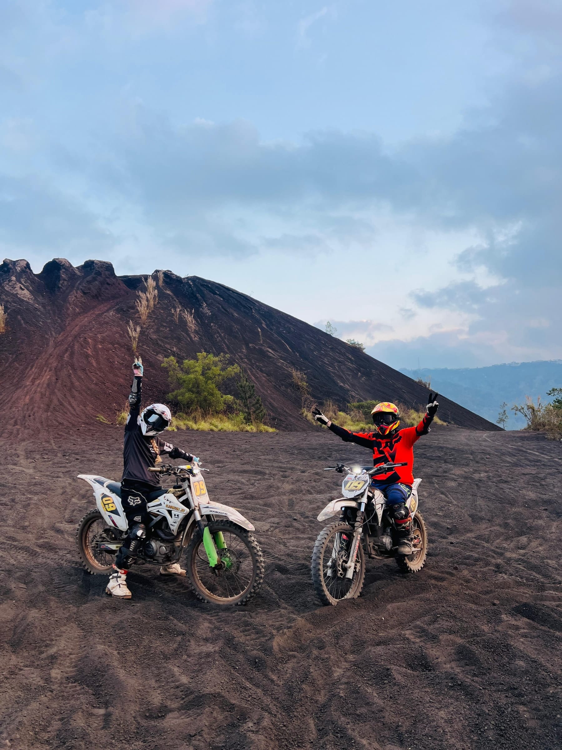Two riders on trail bikes with their arms in the air on the black sand, the Batur cone behind them
