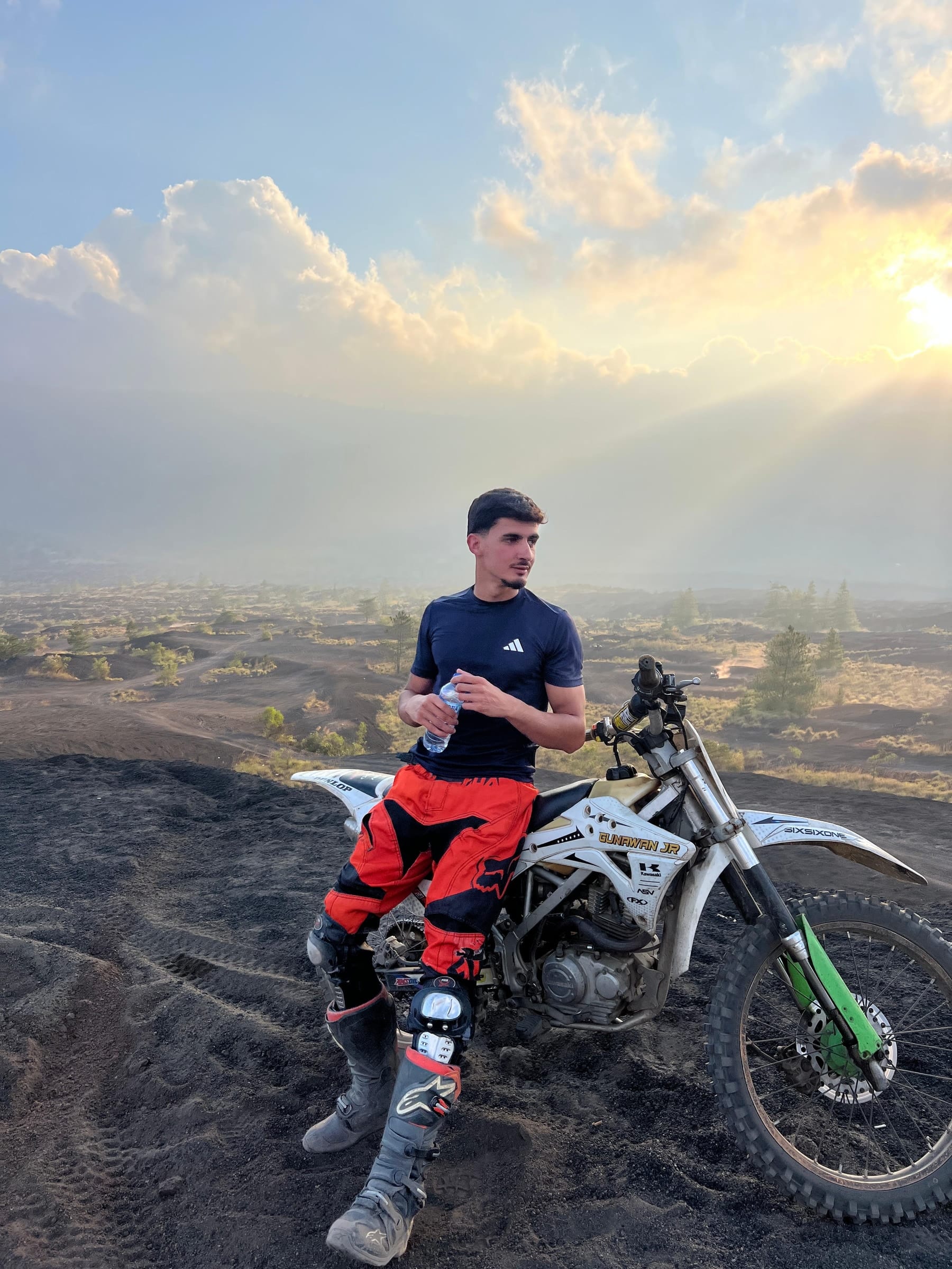 A rider resting against his trail bike on the black sand, sunbeams breaking through the cloud behind him