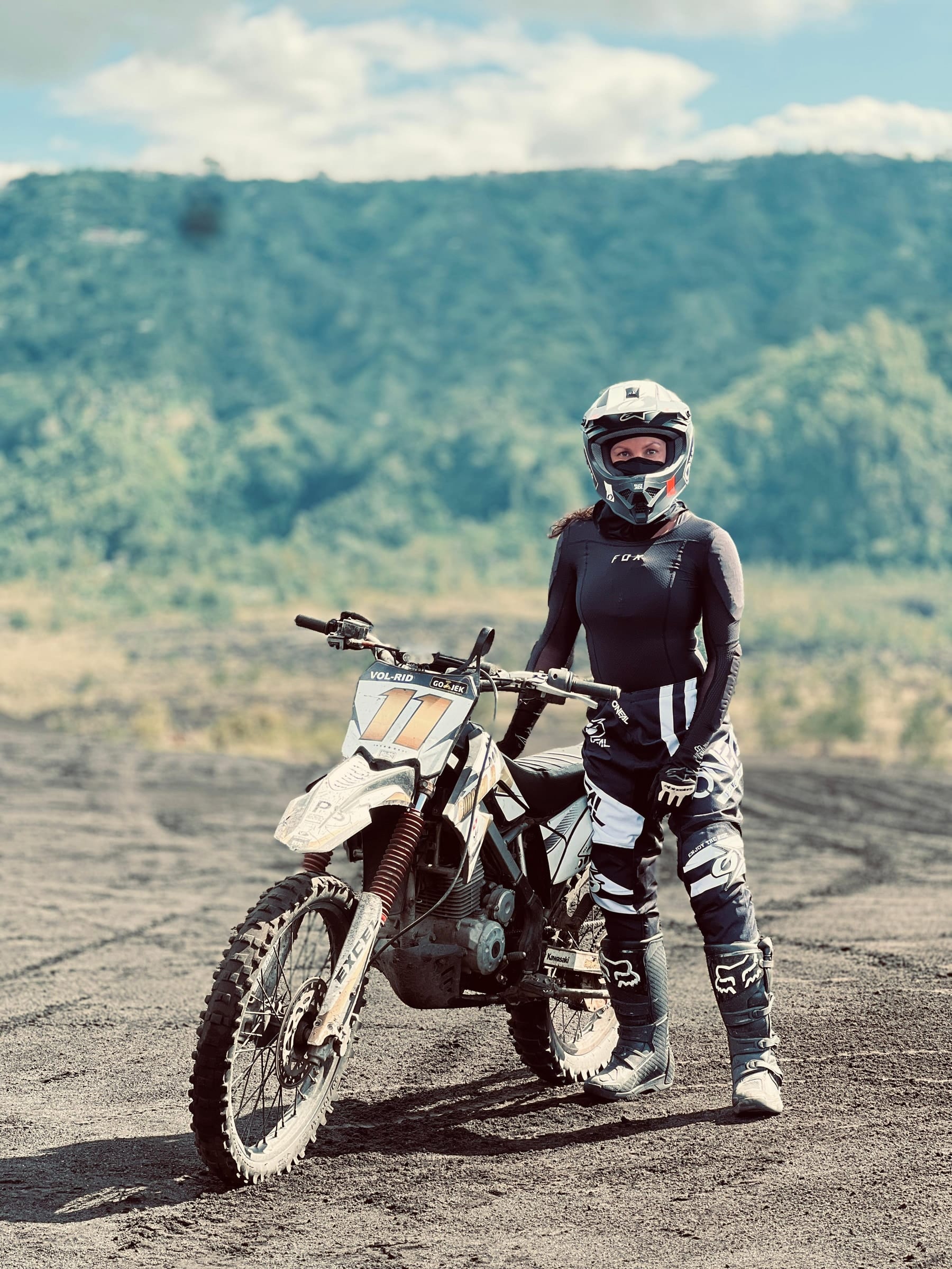 A rider in helmet, gloves and knee braces standing beside her trail bike on the black sand