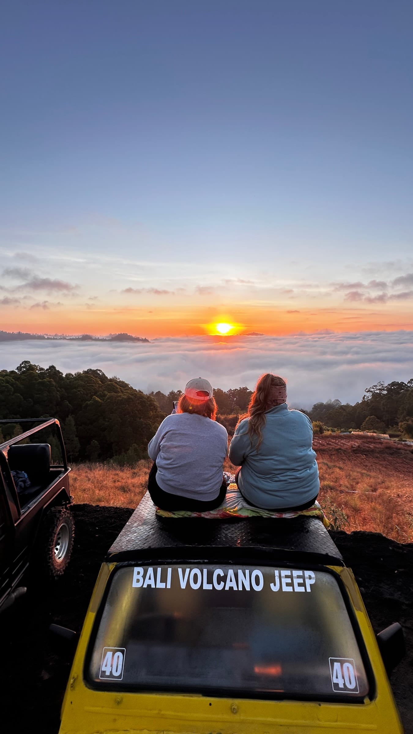 Two guests on a jeep roof watching the sun come up over the clouds