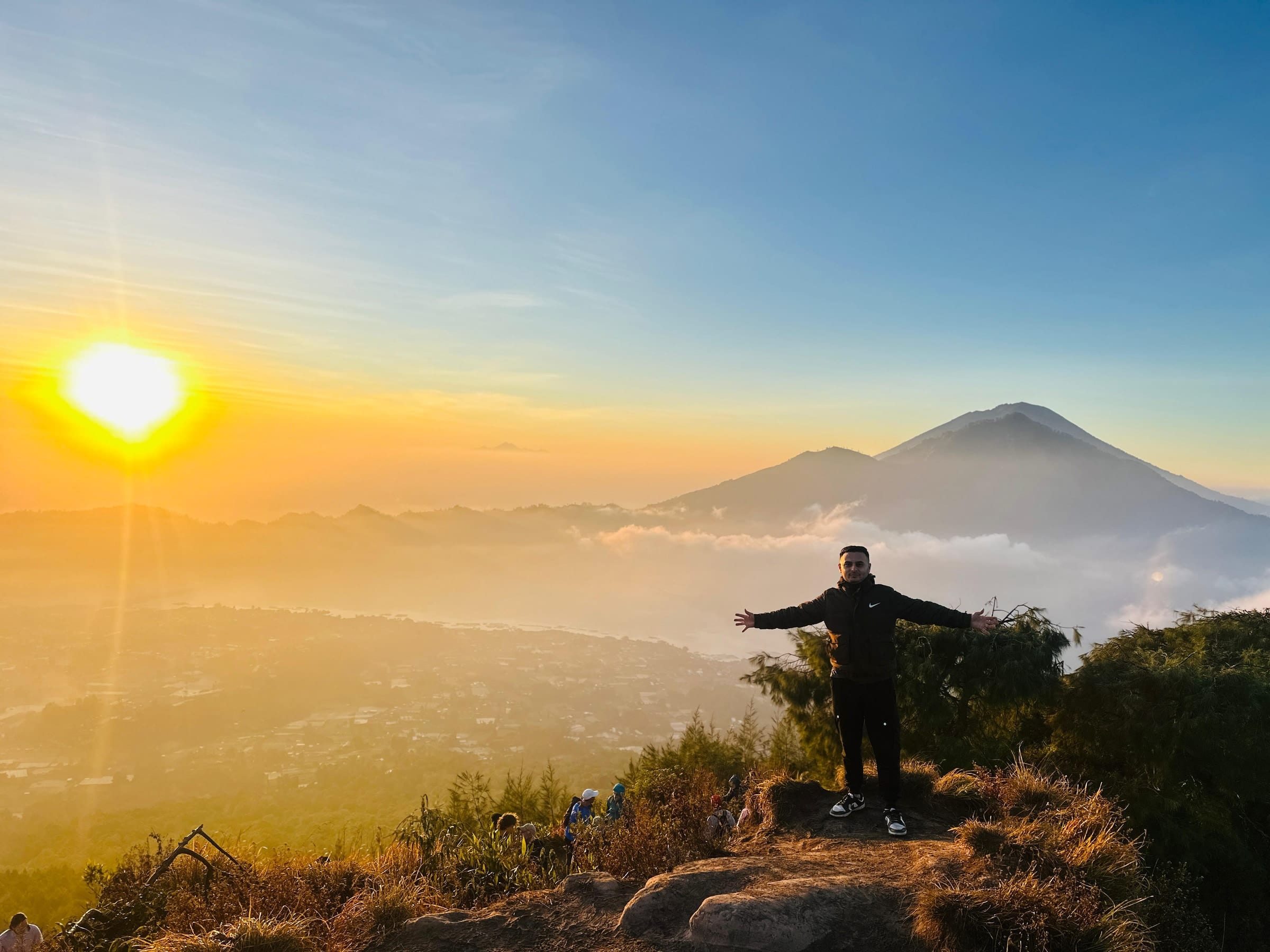 A trekker on the summit with his arms out at sunrise, Mount Agung above a sea of cloud